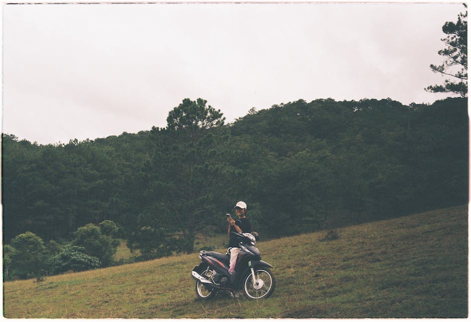A person riding a bicycle in the mountains surrounded by green trees and blue skies. The person is riding uphill on a dirt path, and the bicycle is a mountain bike with wide handlebars and thick tires.