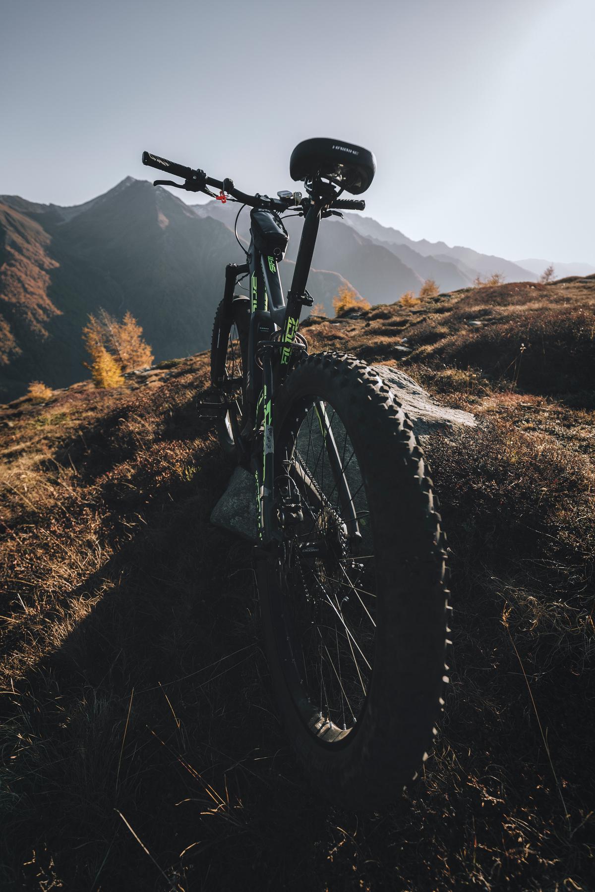 An image of a person on a mountain bike riding down a steep and rocky terrain with trees and mountains in the background. The rider is wearing protective gear, including a helmet and pads, and has a look of focus and determination.